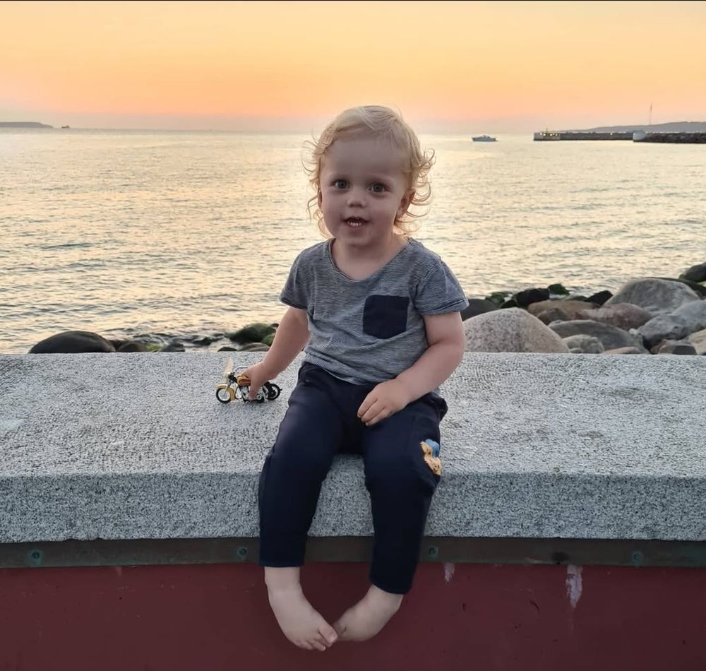 En tvåårig, blond, lockig pojke sitter på en mur. Bakom honom breder havet ut sig i solnedgången, himlen är färgad rosa och orange. Bilden är tagen i Helsingborg.