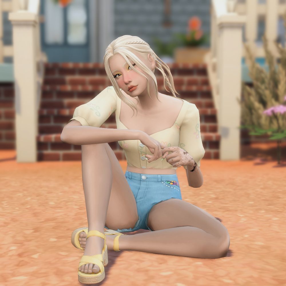 A blonde sim, dressed in a pale yellow cropped top, with light blue denim shorts, and yellow wedged heals. She's sitting on the ground, with one knee up, her arm resting on it. 