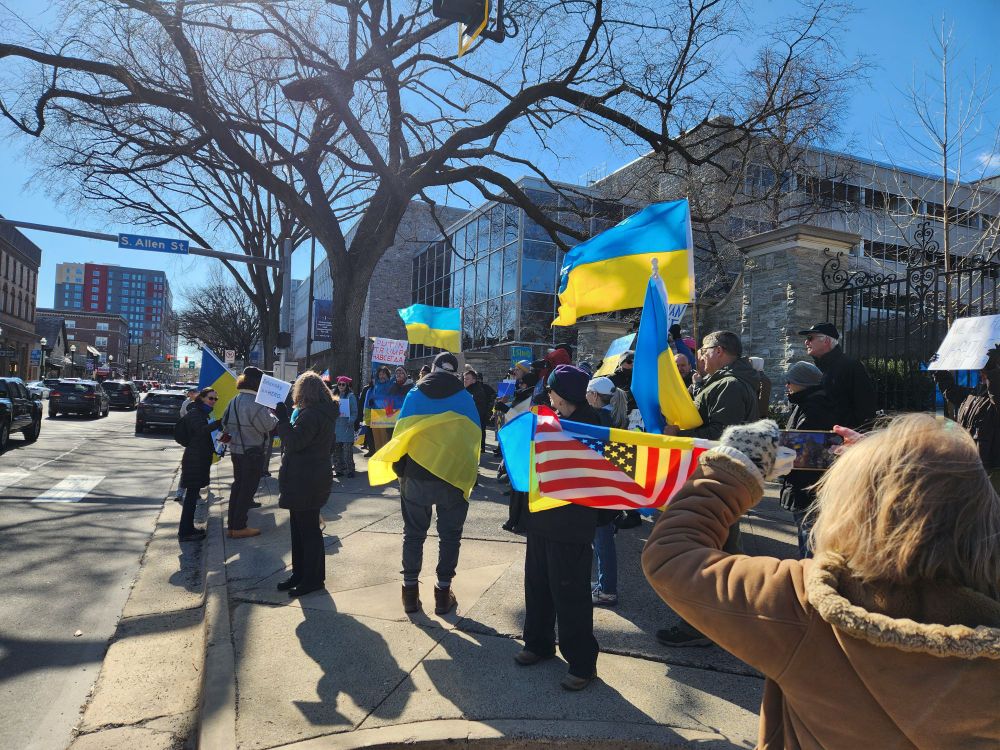 A crowd of people demonstrating holding protest signs and Ukranian and American flags.