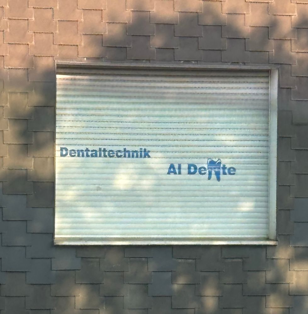 Fenster mit heruntergelassenem weißen Rollladen, auf dem in blauer Schrift ‘Dentaltechnik’ und ‘Al Dente’ mit einem stilisierten Zahnlogo steht. Das Gebäude ist mit dunkelgrauen, schuppenförmigen Fassadenplatten verkleidet. Licht und Schatten von Bäumen spiegeln sich auf dem Rollladen wider.