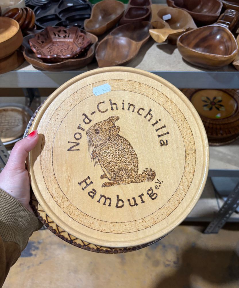 Eine runde Holzplatte mit einem eingebrannten Motiv eines Chinchillas und der Aufschrift “Nord-Chinchilla Hamburg e.V.”. Die Platte wird von einer Hand gehalten und steht zum Verkauf für 2,00 €. Im Hintergrund sind verschiedene Holzschalen und -gefäße in einem Regal zu sehen.