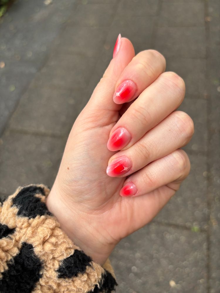 Eine Hand mit rotem Nail-Art-Design vor einem unscharfen Gehweg-Hintergrund. Die Nägel sind mandelförmig gefeilt und mit einem auffälligen roten Farbverlauf und glänzendem Finish gestaltet