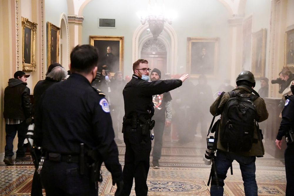 A photograph of a stand-off between Capitol Hill security and January 6 insurrectionists. Tear gas and smoke fills the room.