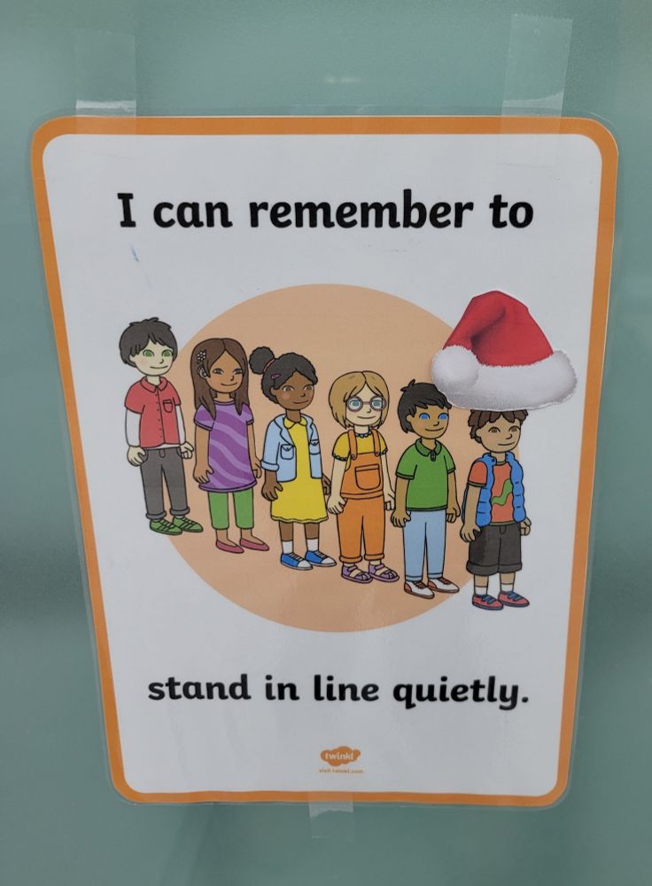 a sign with children standing in a line. the child in front has a Santa hat on. 
