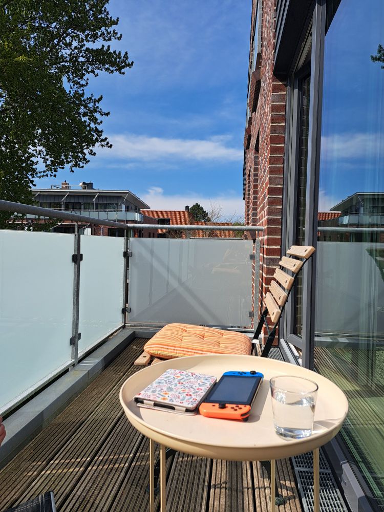 Kindle, Switch und ein Glas Wasser auf einem Balkontisch, im Hintergrund blauer Himmel, kaum bewölkt