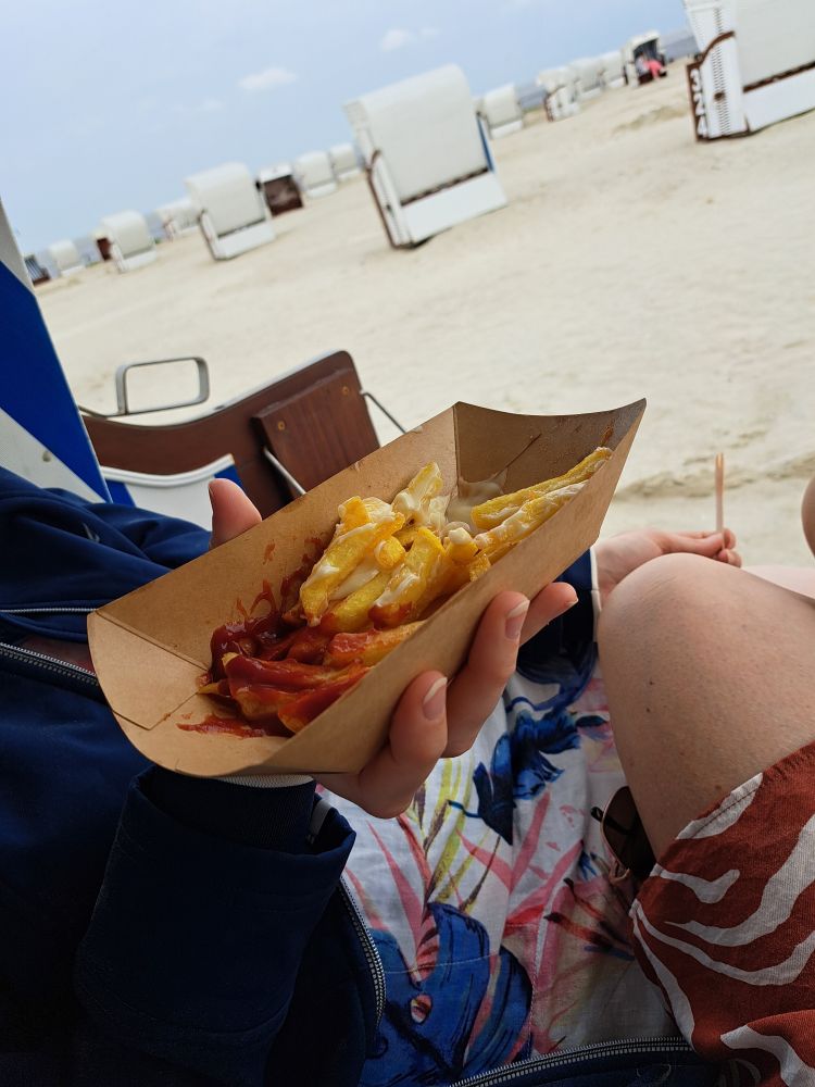 Pommes rotweiß in Pappschale, gehalten von einer Hand, am Strand