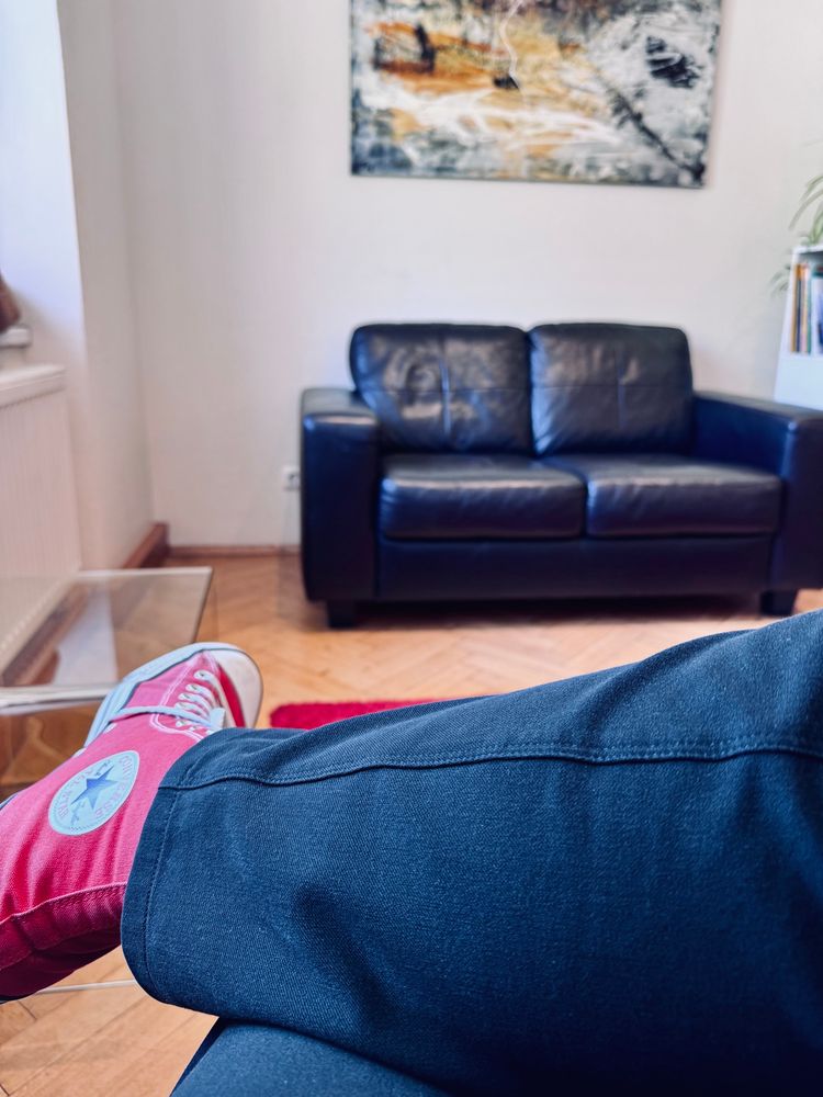 Ein Wartezimmer. Man sieht im Hintergrund moderne Kunst und einen schwarze Ledercouch. Ansonsten sieht man den Holzboden und meine Füße. Ich trage eine schwarze Stoffhose und rote Chucks.