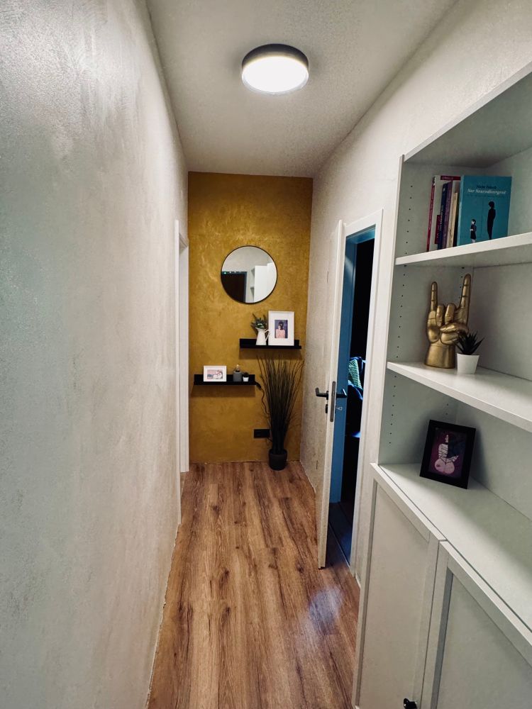 Ein kleiner Flur. Die Wand auf der Stirnseite ist Goldfarben mit zwei schwarzen Regalbrettern, Spiegel und und zwei Bilder . Auf der rechten Seite steht noch ein Bücherregal mit ein paar Büchern, einem Bild und einer goldenen Hand, die einen Mano cornuta zeigt.