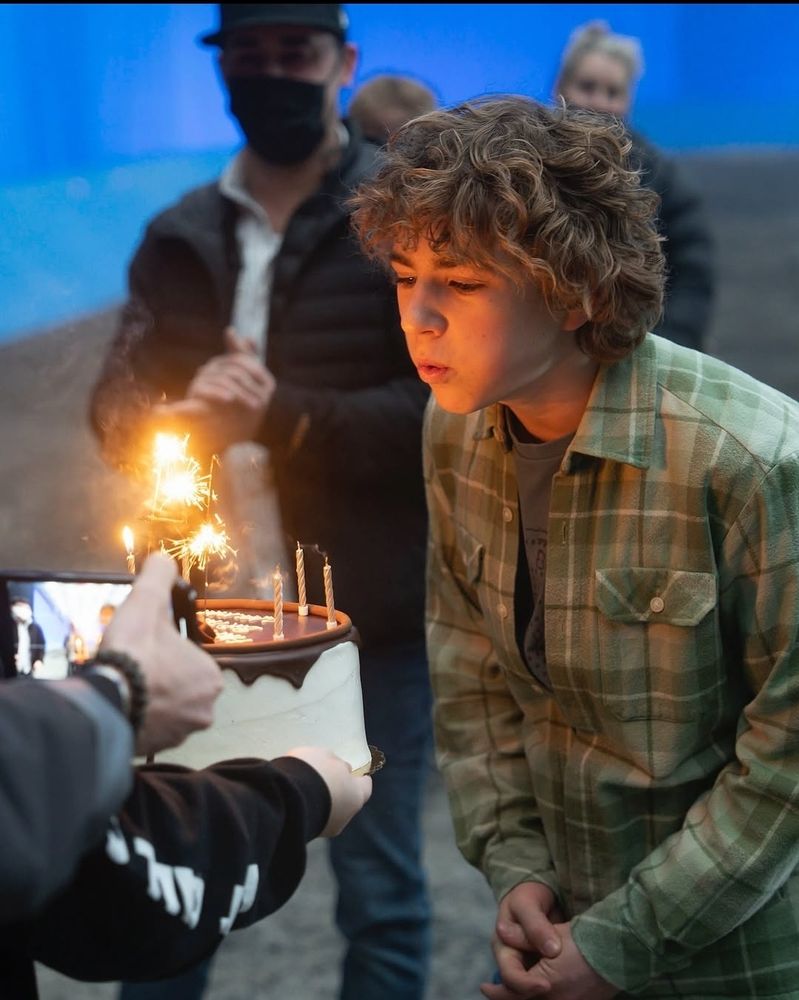 Walker blowing candles on his 14th birthday, on the set of PJO S1, behind him a few people, a blue screen, and a bunch of sand