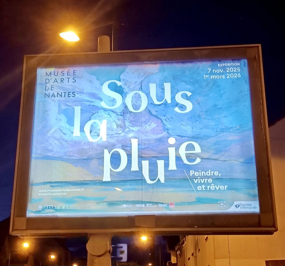 Panneau publicitaire lumineux de nuit dans une rue illuminée : affiche d'une expo appelée "Sous la pluie" - "Peindre, vivre et rêver", une peinture d'un paysage de plaine sous un ciel nuageux chargé, au Musée d'Arts de Nantes, du 7 Nov. 2025 au 1er Mars 2026.
