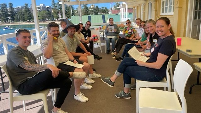 Pic of Nat and participants on the Nov 24 Resuscitology course. One holds a banana as a phone; one has a rubber chicken; one is wearing a wizard hat; one holds a picture of a bee; one has a Rubik’s cube; one has a bougie; and all are smiling on a gorgeous sunny day in Manly. John Hinds’ motorbike helmet has a chair of its own.