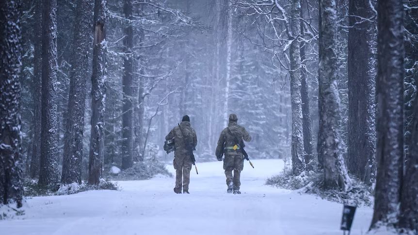 Deux personnes en tenue de camouflage marchant dans une forêt enneigée sur un chemin couvert de neige.