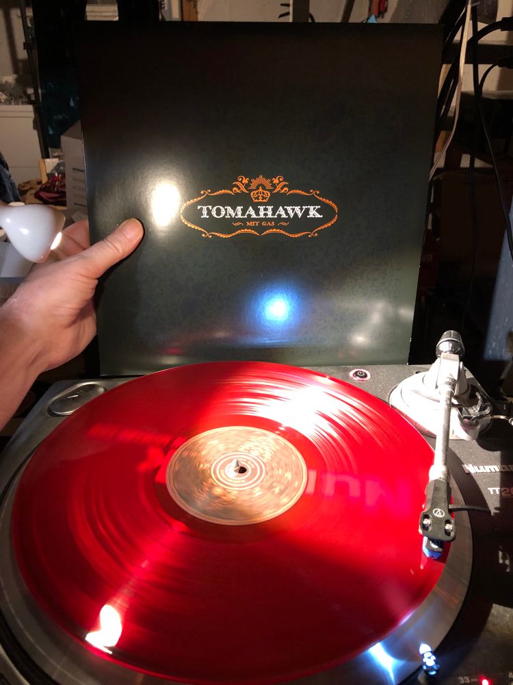 Candy-apple red record spins on the aluminium/steel wheel while a hand holds its black jacket behind the turntable. There's some coppery flourishy decorations around a fancy, allcaps white font that reads TOMAHAWK and, in the same font but smaller and copper below it, MIT GAS. Fukken beauty 🤘❤️‍🔥⛽️