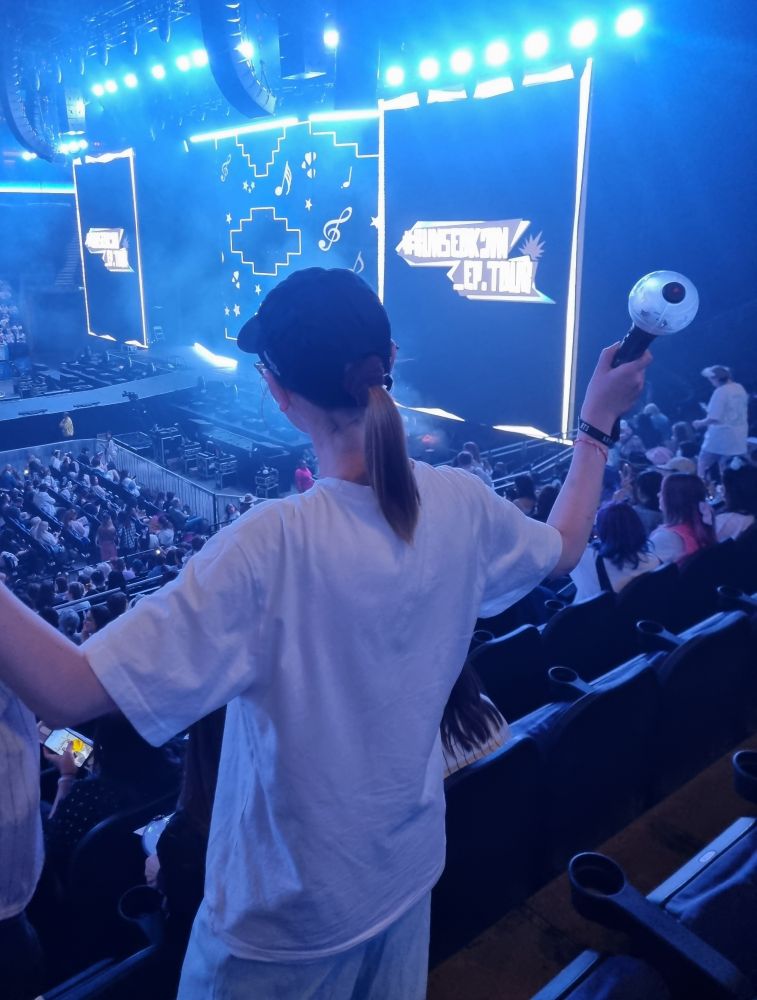 Chrissy, a thin white woman, standing inside the O2 for Run:Seokjin ep. Tour in London. Her back is facing the camera and she holds an ARMY bomb in her right hand. She wears a black ball cap and her hair is tied up in a ponytail. She also wears a white oversized T-shirt and wide leg jeans. 