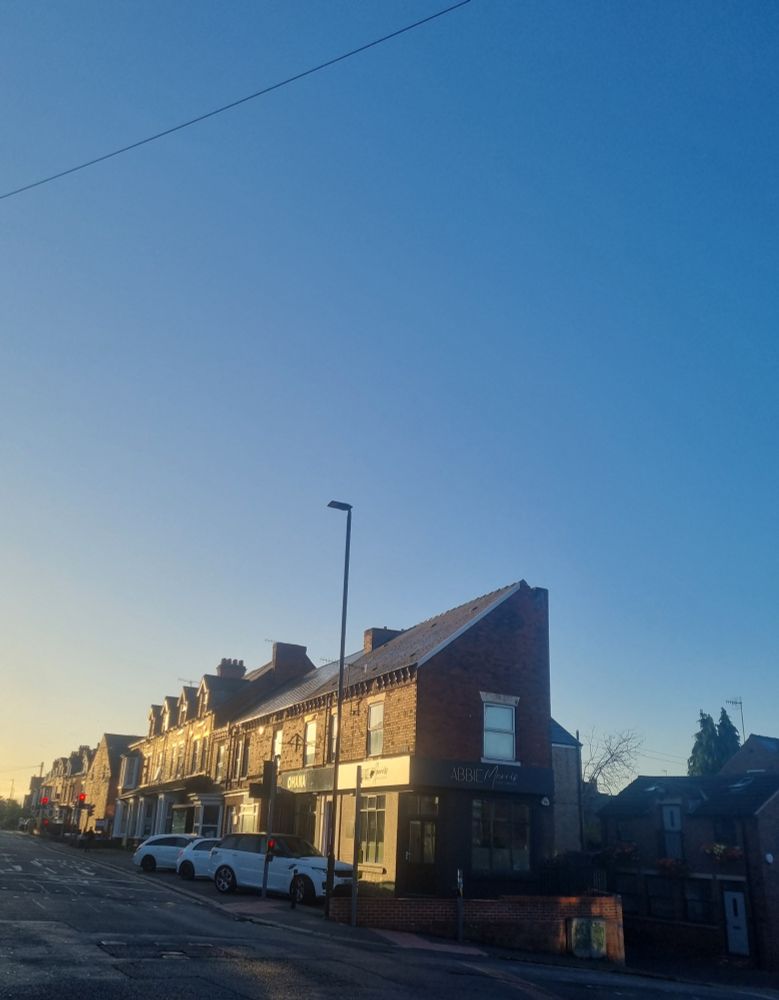 Blue morning skies over a row of houses. There is an electric wire spanning from the top left corner to the right. The houses are bathed in golden morning light, and the sky is blue and cloudless.