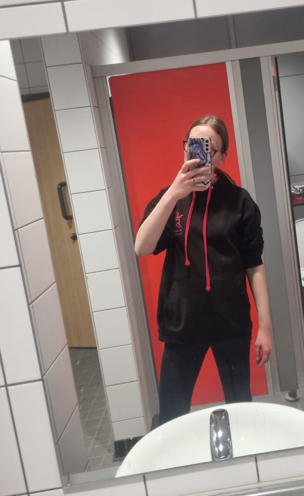 Mirror selfie in a gym bathroom. Chrissy, a thin white woman, is standing in front of a red door. She holds her phone up so that it covers her face. She wears a black hoodie with red drawstrings and a red logo, and black leggings. 