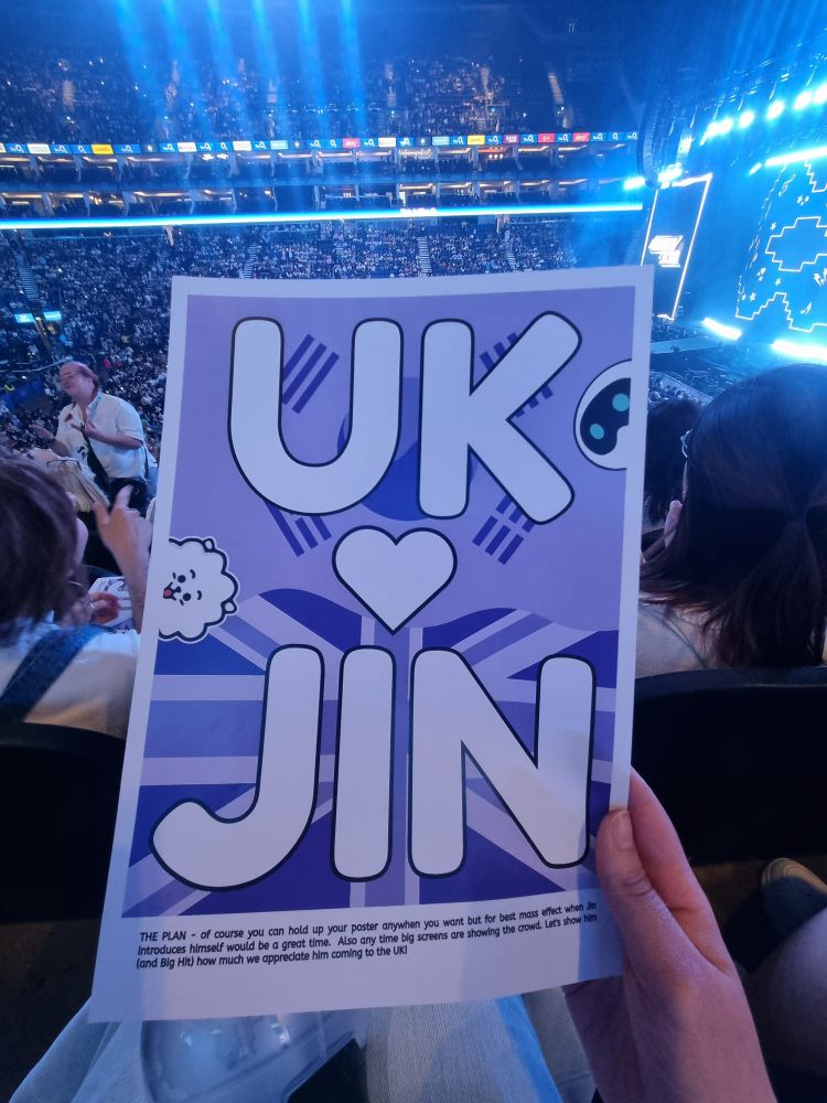 Chrissy holding the "UK ♡ Jin" banner for Jin in London, day one. The banner is A4, it has a purple Korean flag & Union Jack in the background. The slogan is written in bold white letters, and RJ & Wootteo peek out cutely from each side of the banner. 