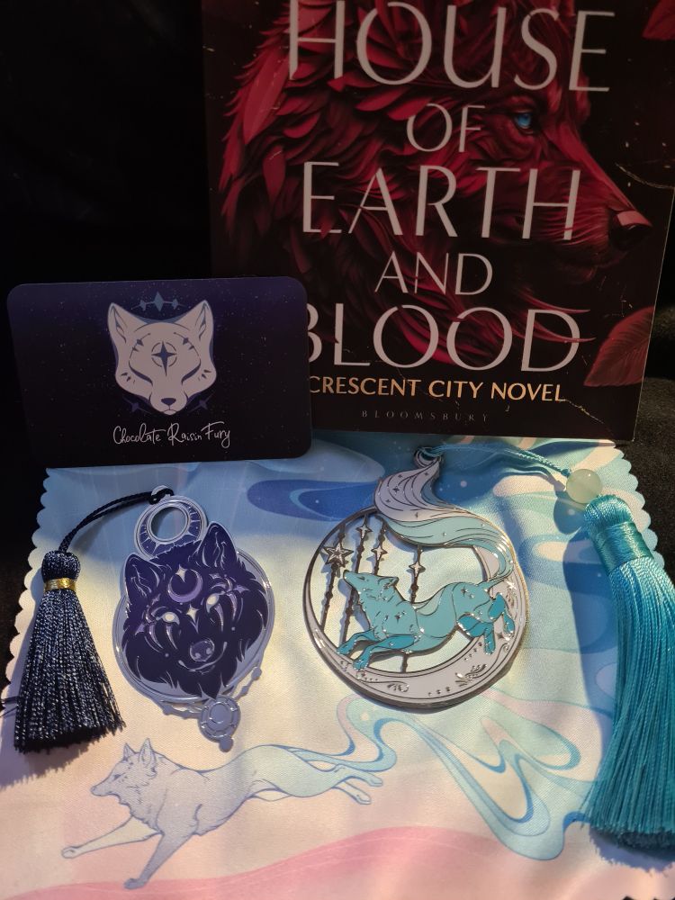 Two metal bookmarks, one of a wolfs head in purple and silver, the other a fox leaping on a cresent moon with stars in light blue and white, on a lense cloth with a book and business card behind