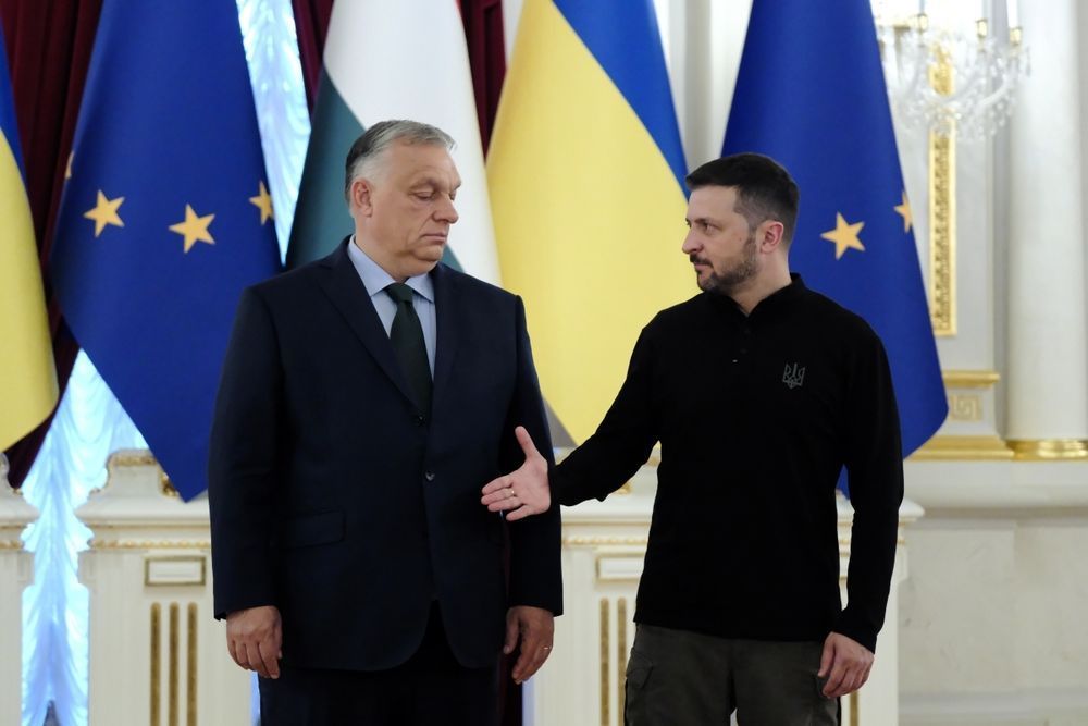 Zelenskyi reicht Orban die Hand hin, Orban schaut mit betont verächtlichem Gesicht darauf hinunter. Im Hintergrund Fahnen der EU und der Ukraine.