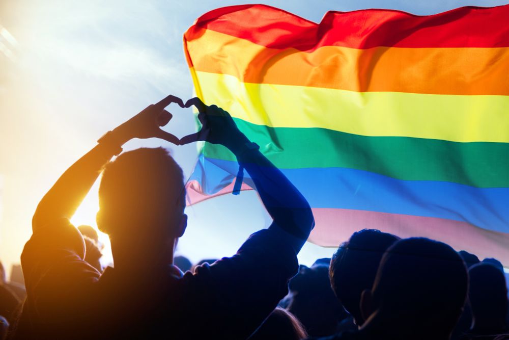 A person standing in shadow making a heart with their hands over their head as a Pride flag flies in the background