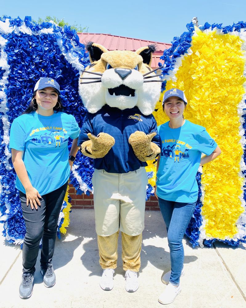 Linda Chang and a colleague take a photo with Rufus Bobcat in front of a decorative background with the UC Merced "M" logo during a bright sunny day.