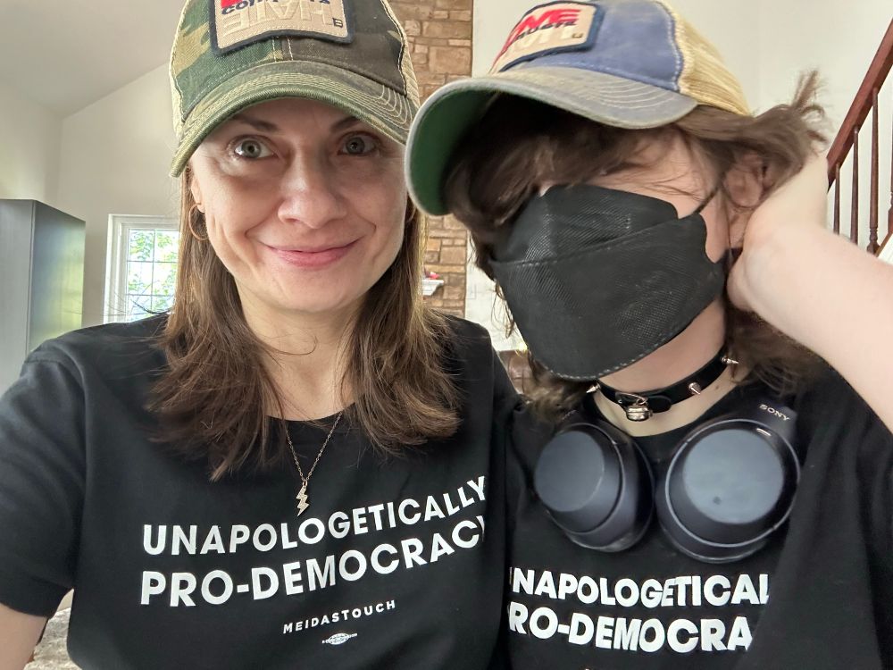 Two folks, a mom and kiddo, one in mask, with T-shirts “unapologetically pro-democracy” and MeidasTouch on black shirt 
