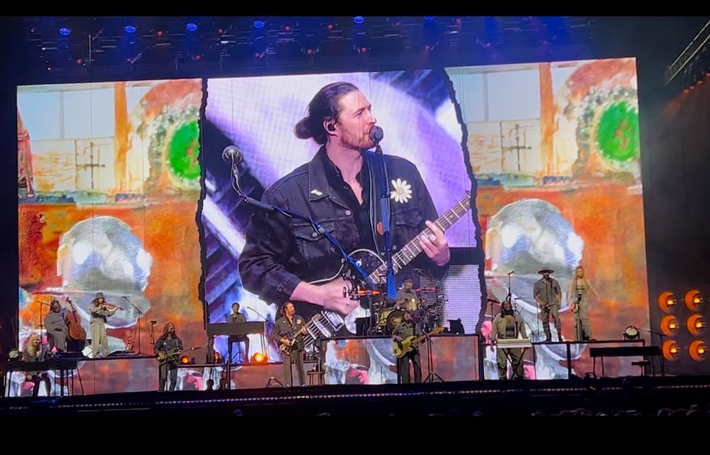 Hozier playing guitar and singing on stage with a close up of him in denim jacket with daisy pin for peace