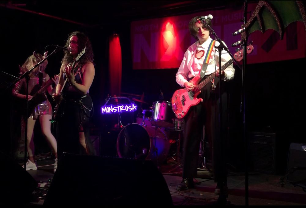 Musicians standing on a stage. On the left is a woman with blonde hair dressed in a pink knight outfit. She is holding a black guitar. In the middle of the stage is a woman with shoulder length, curly black hair. She is holding a purplish-blue guitar and is singing into a microphone on a microphone stand. On the right side of the stage is a woman with short brown hair in a white dress shirt. She is holding a pink bass guitar. At the back of stage is a drum kit. There is a purple neon sign near the kit. It reads: "MONSTROSA."