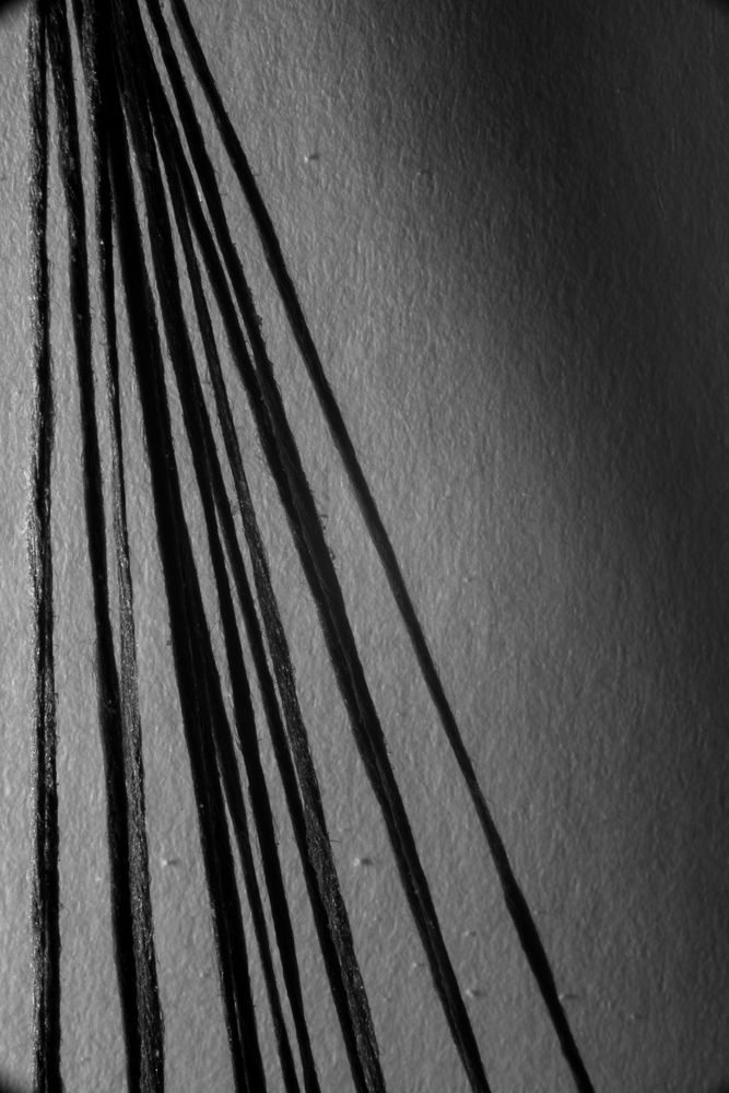 Black and white photo of black thread drawing vertical lines that gradually increase in the angles that separate them. They mix together with shadows that they themselves cast on a fibrous sheet of paper. A non-de script shadow looms over the top right portion of the image. 

Personal note: I didn't quite nail the effect I was going for with this one but I'm not too displeased with the result.