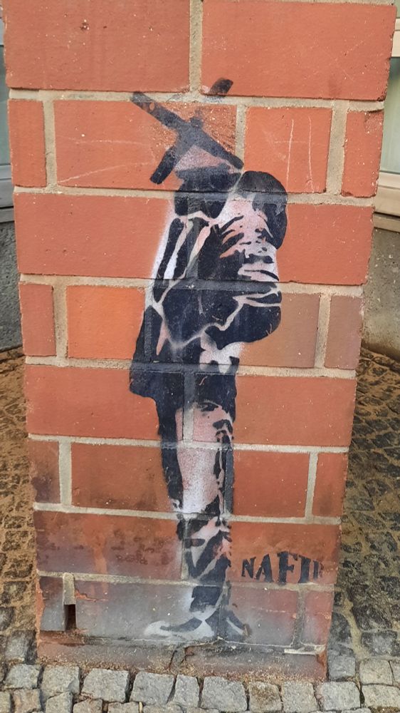 Auf einem Pfeiler gemalt eine Person in schwarzweiß, das Gesicht ist nicht zu erkennen und mit einem schwarzen Kreuz markiert. 