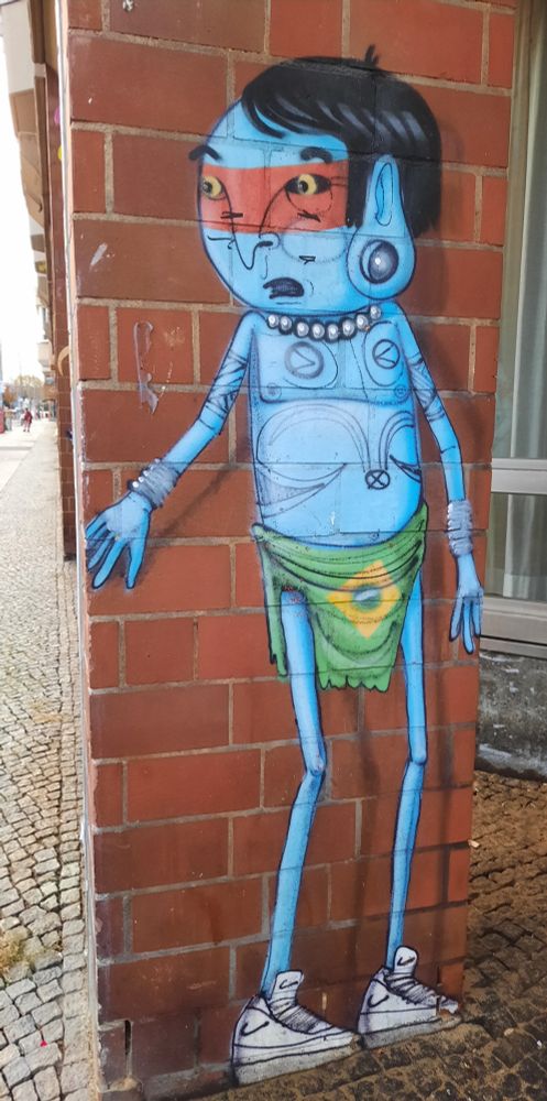 Auf einem Pfeiler gemalt ein blaues Männchen, nur mit einer brasilianischen Flagge um die Hüften bekleidet. Es guckt hinter sich.