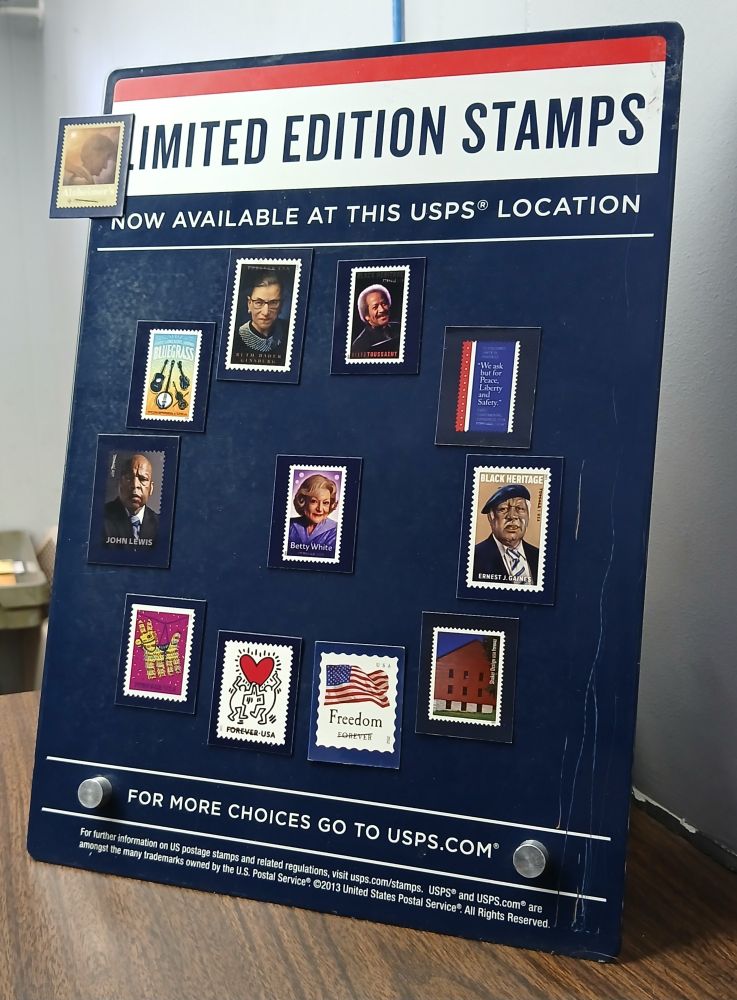 The new Betty White stamp is surrounded by other commemorative stamps including John Lewis, RBG, the art of Keith Haring and regular flag stamps and more, set on a USPS stand up placard on a USPS counter, titled "Limited Edition Stamps" and "for more choices go to usps.com" 🕯