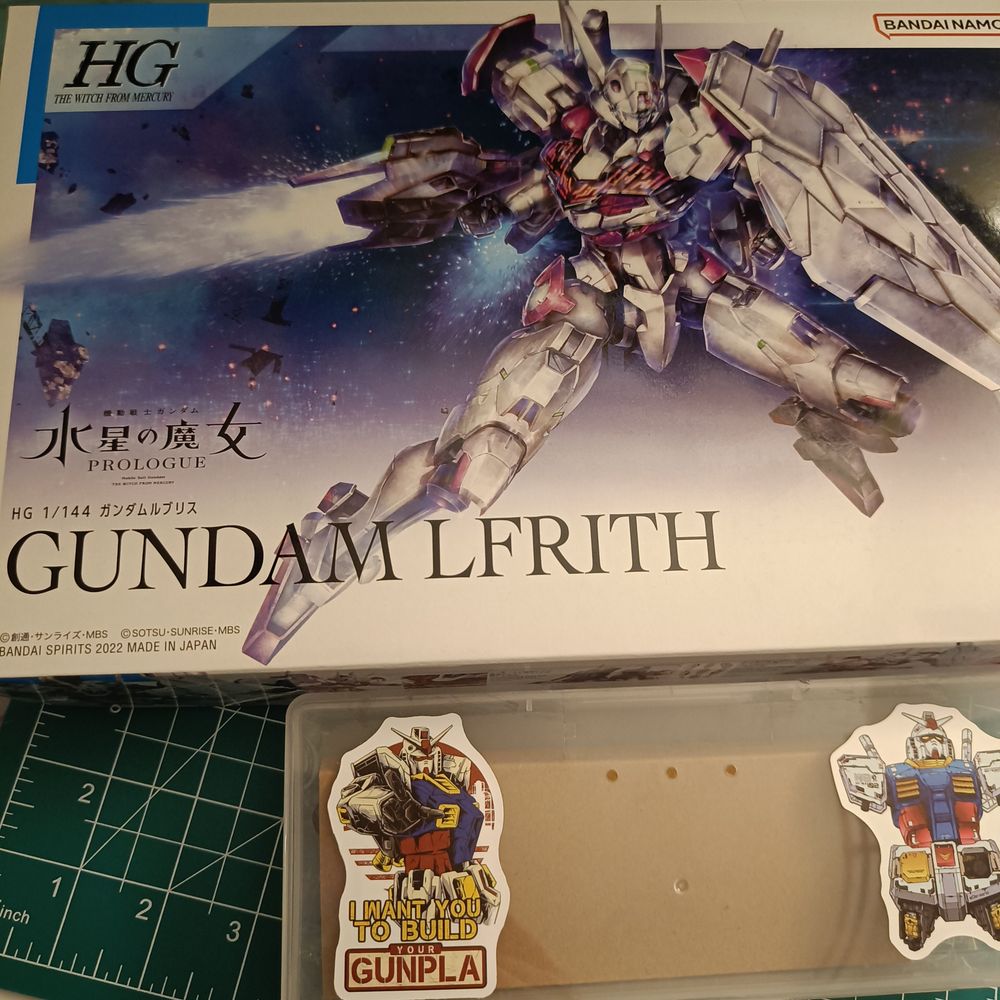 A photo of a gunpla model kit, a box of tools with stickers and a cutting mat. The model kit is the HG Gundam Lfrith from prologue of The Witch From Mercury anime.