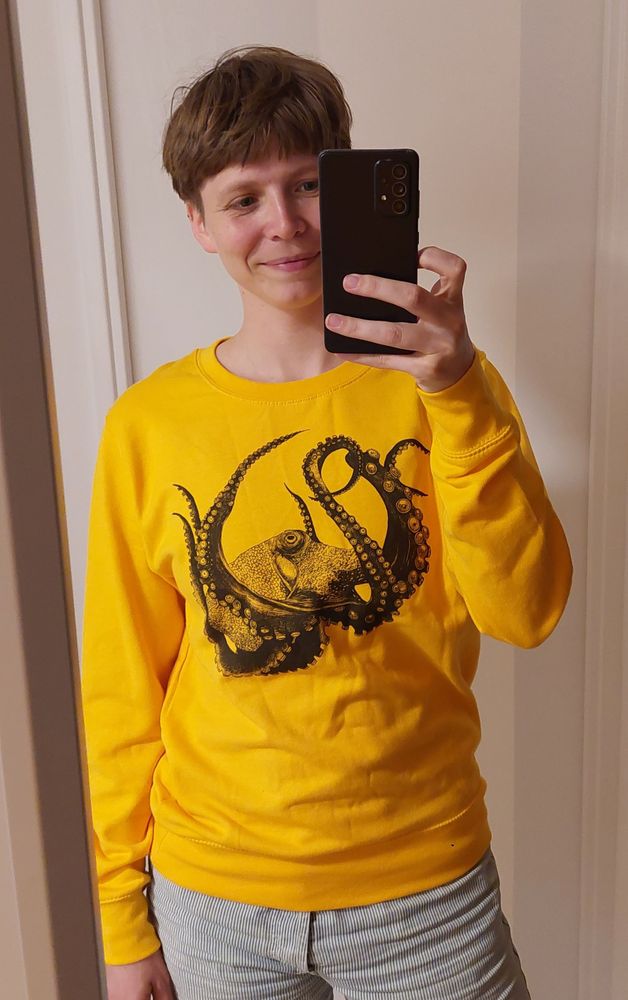 Mensch mit gelbem Pulli mit großer schwarzer Krake darauf, der in die Kamera lächelt