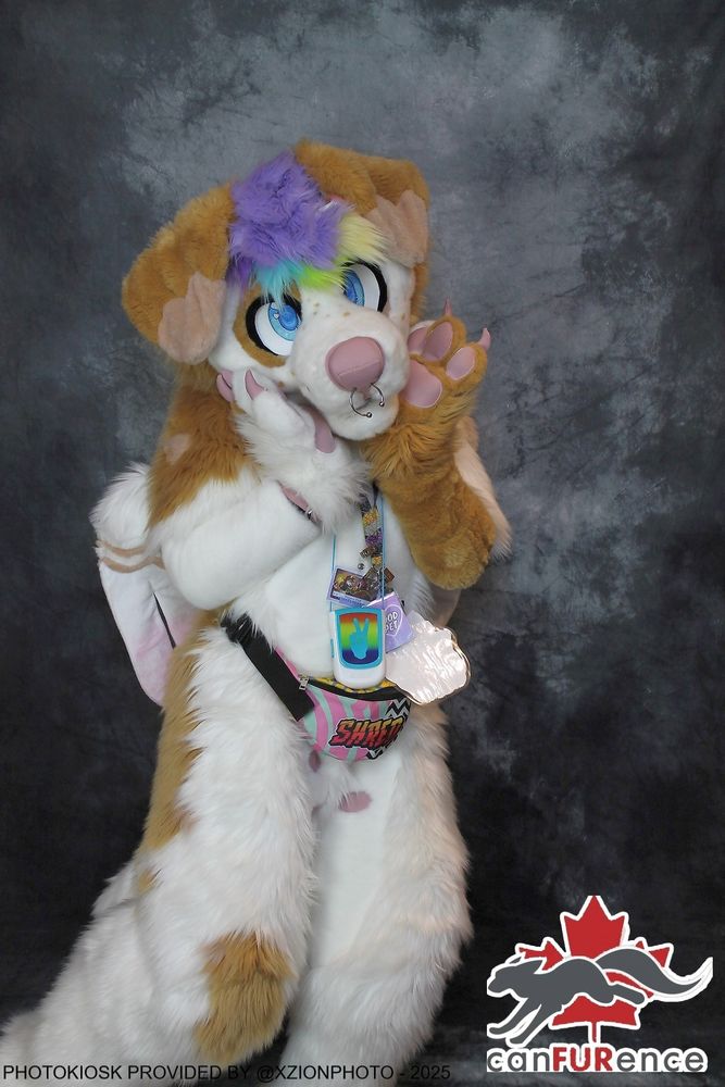 A white dog fursuit with brown spots and lilac hair with rainbow accents. He is cupping his face in his paws and looking at the camera
