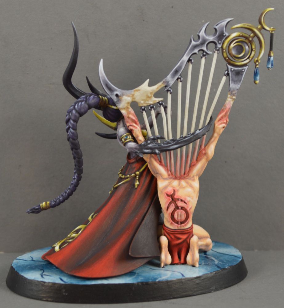 Painted miniature of a non-human but humanoid demon playing a harp that is sprouting from the back of a human. The human is on his knees, bald, and wearing a red loincloth. The demon has pale purplish skin, dark horns, and is wearing a fancy red dress with the symbol of Slaanesh on it in gold. The back view shows that the symbol has also been carved into the human’s back. This is why we do not bend an inch for Chaos. It will end badly.