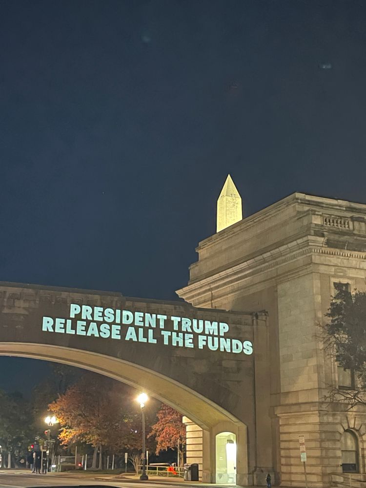 Projection on USDA building with Washington Monument in the background. Projection reads “President Trump release all the funds”