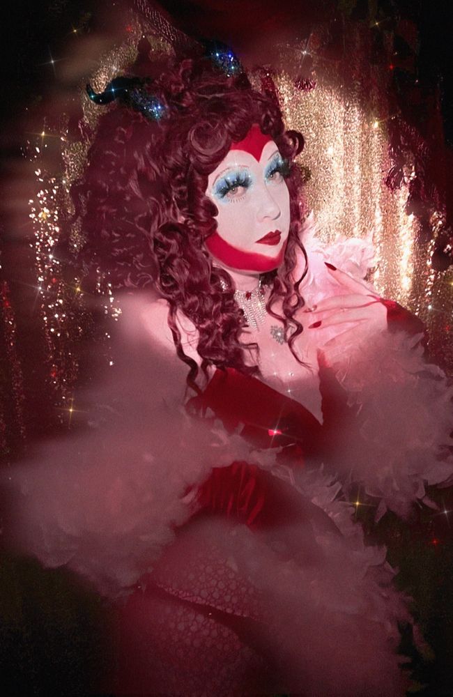 Daisy poses in a drag look inspired by Chappell Roan’s my kink is karma music video look. They have a big white heart on their face outlined in red with giant lashes and blue eyeshadow. They are wearing a red getup and pink feather boa with black bedazzled horns in their giant red wig