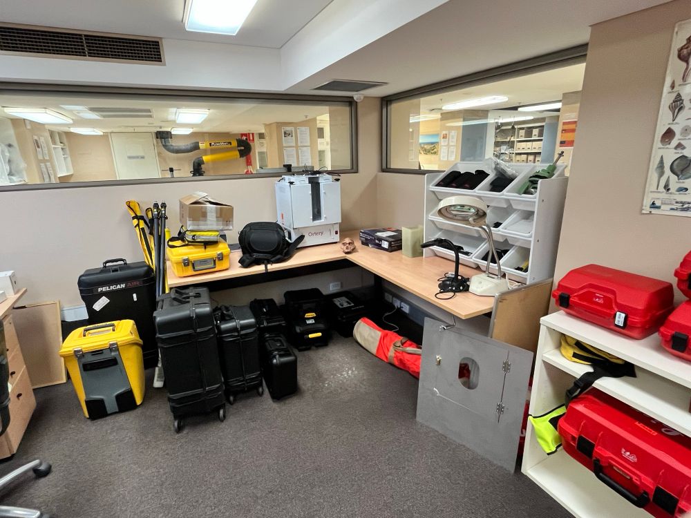 The Archaeology Hub office showing part our collection of surveying gear. Multiple protective cases in the background and on the desk, along with a light box and two magnifying lamps. The model skull of an Australopithecus is also on the centre of the desk.