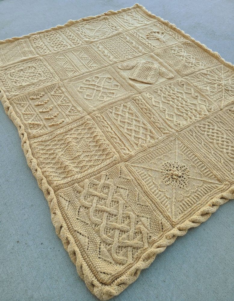 A pale yellow blanket, made with squares of different knitted cable patterns. It's spread out on concrete.