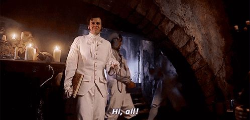 a man in a white suit stands in a dark room and says hi all