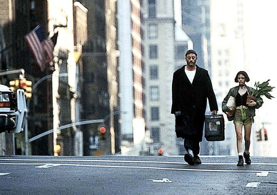 A still from the film Leon: The Professional. The characters Leon and Mathilda walk down the middle of an avenue in New York City. Leon wears a long black coat with a white shirt underneath and holds a large briefcase. Mathilda wears a green jacket and shorts with a black top and holds a midsize green plant.