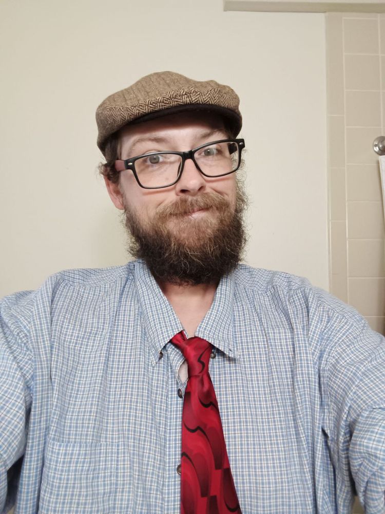 Me wearing a blue checkered dress shirt, a somewhat loose fitting red and black tie, a tan colored newsboy cap, and black glasses with red earpieces. I have a pretty bushy beard and I'm smiling with my mouth closed.