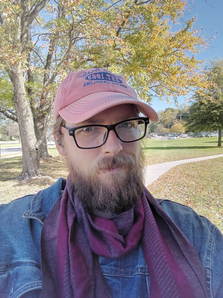 A selfie of me, Ben. Denim jacket, burgundy colored scarf, orange hat, black and red glasses and busy brown beard
