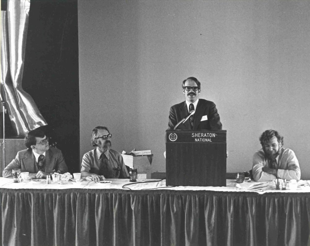 Caption reads “Jim Fry (University of Michigan) speaks to the audience while James Rothnie (Computer Corporation of America), Edgar H. Sibley (University of Maryland), and John Gosden (Equitable Life Assurance Society) look on at the ACM SIGMOD Conference in Washington, D.C. during June 1976.”