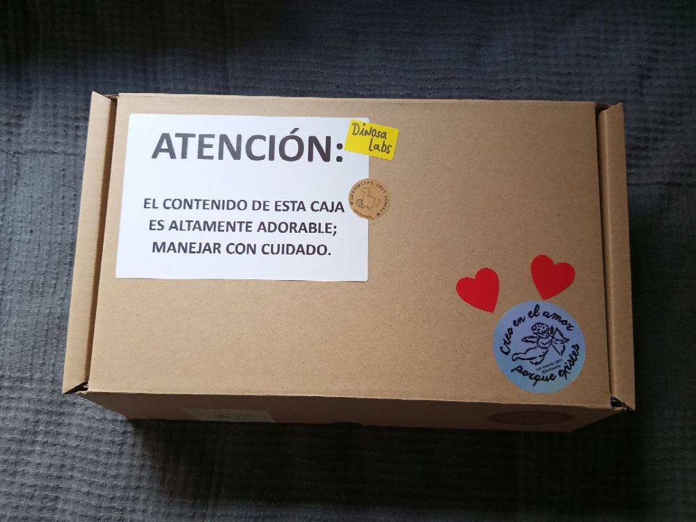 La cajita cerrada, con una etiqueta que reza : "ATENCION: El contenido de esta caja es altamente adorable; manejar con cuidado", lleva además dos pegatinas de corazones y una con cupido que dice "Creo en el amor porque existes"