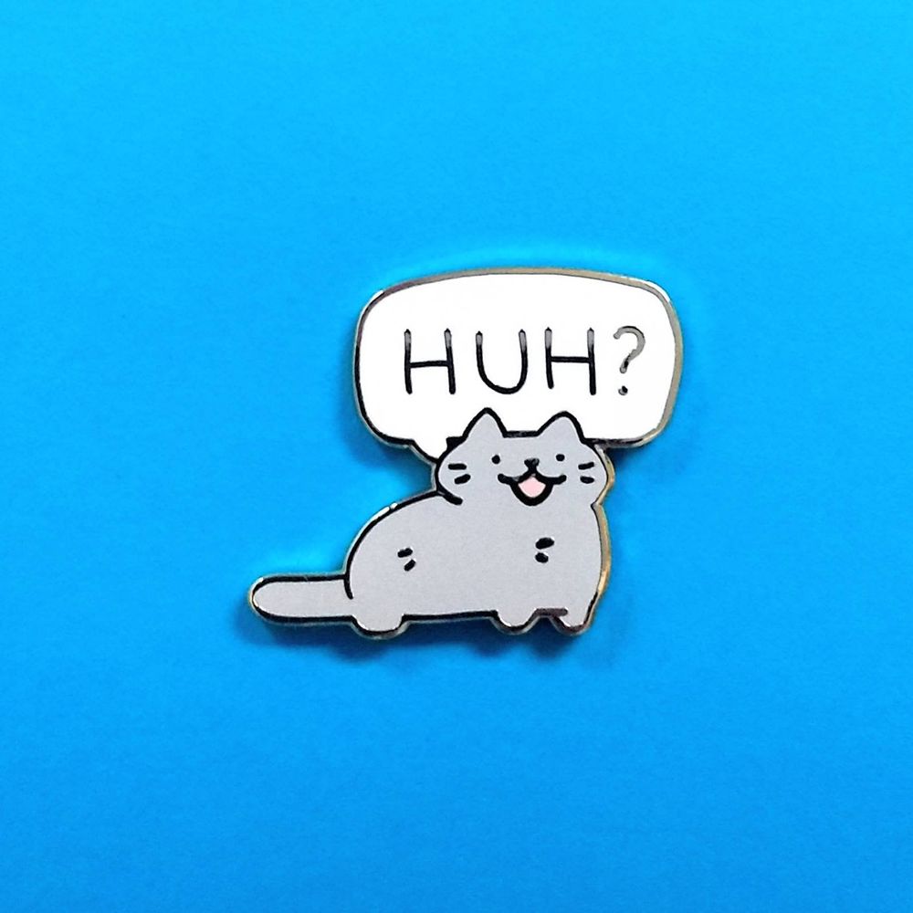 enamel pin of a gray cat saying "HUH?"