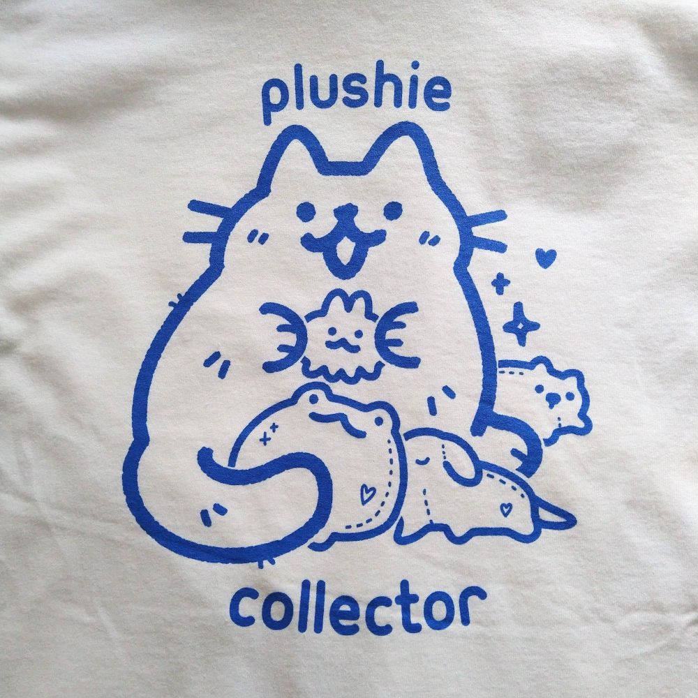 White tee with blue ink printed graphic of a cat drawn in a cute style holding various plushies. text "plushie collector"