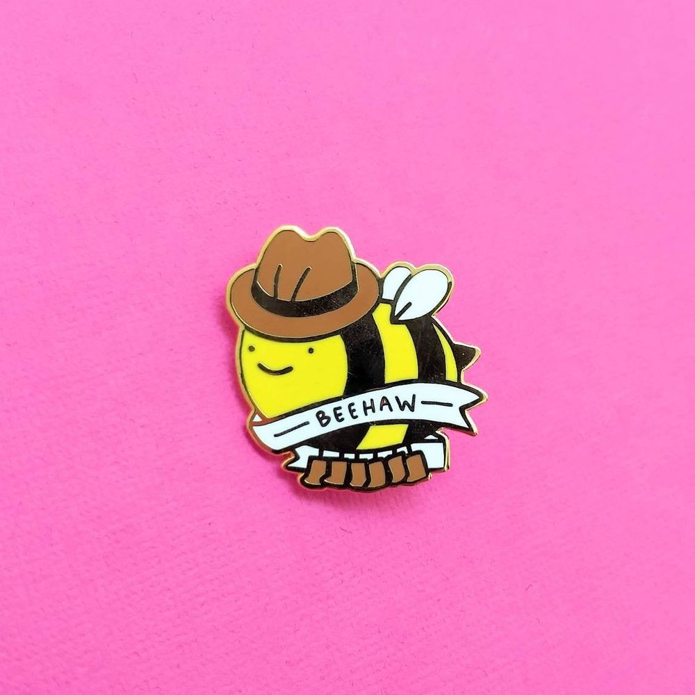 enamel pin of a bee wearing a cowboy hat and six cowboy boots. text reads "BEEHAW"