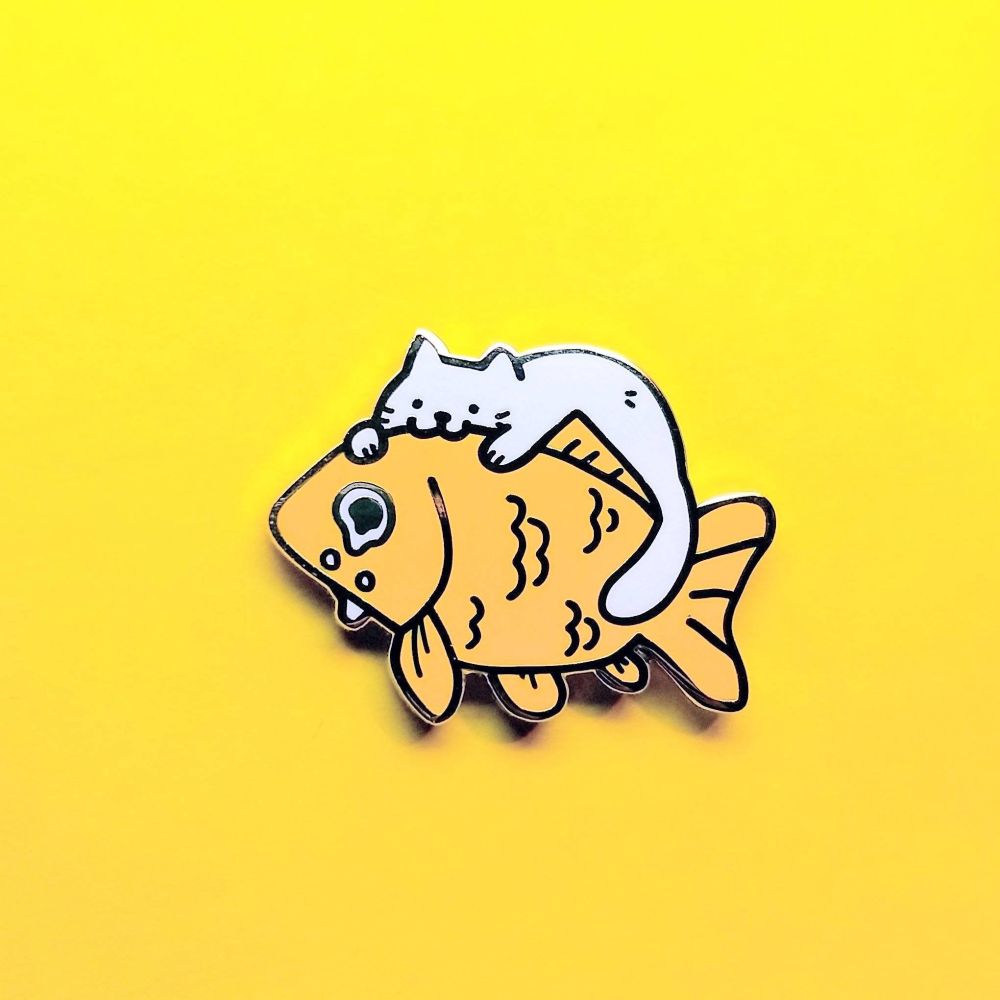 enamel pin of a white cat perched on top of a large crying goldfish. the cat is chomping the fish's head